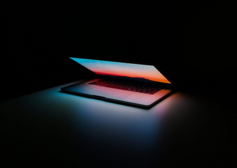 Open laptop in dark setting with vibrant screen displaying blue-to-orange gradient wallpaper, casting colorful glow on surroundings.