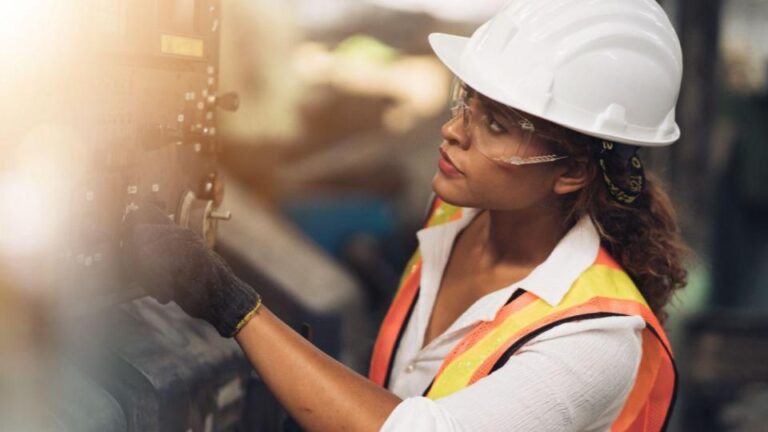 Female engineer in white hard hat and safety vest operating industrial equipment, wearing protective glasses