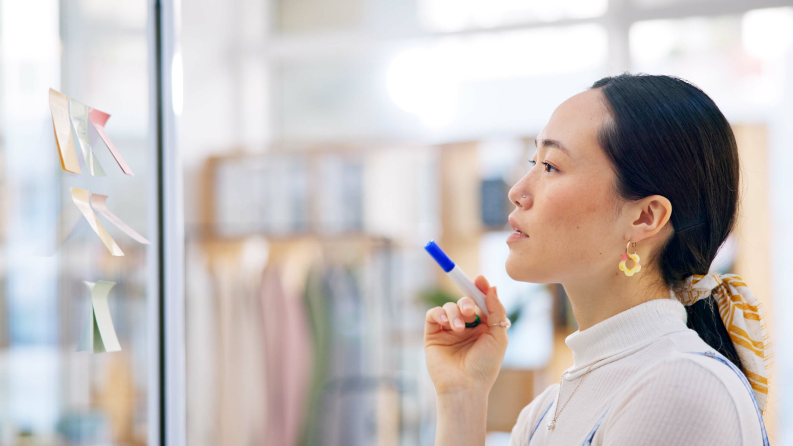 Woman with hair tied back looking at sticky notes on glass wall, holding marker, in bright office environment