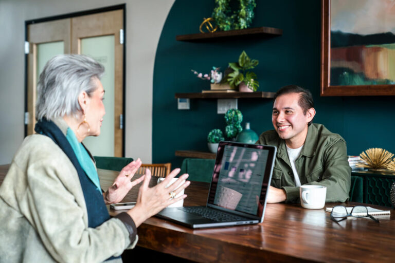 Woman with silver hair in beige blazer gestures while speaking to smiling man in green jacket. Laptop on wooden table. Teal wall with plants and artwork.