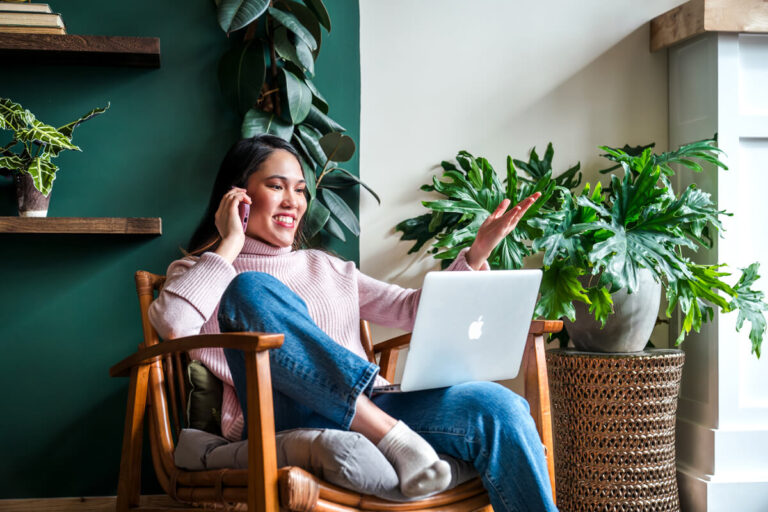 Woman in pink sweater smiling during video call on MacBook, relaxing in wooden chair surrounded by houseplants against teal wall.