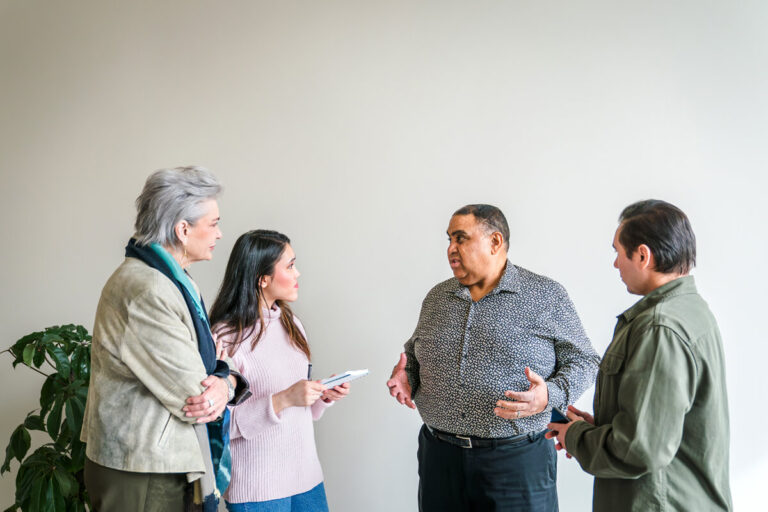Four diverse professionals in conversation against white wall; woman with silver hair in beige jacket, woman in pink sweater, man in patterned shirt gesturing.