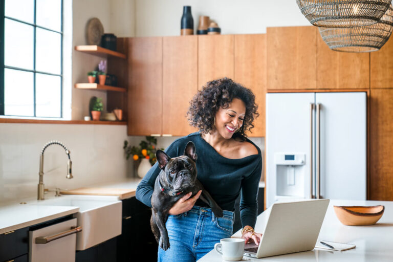 Smiling woman with curly hair holding French bulldog while using laptop in modern kitchen with wooden cabinets, farmhouse sink and white counters.