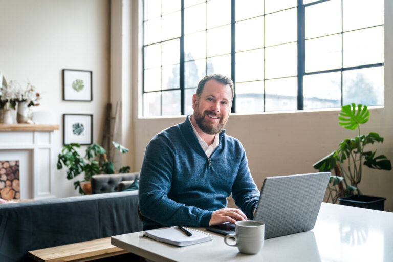 A bearded man in blue sweater smiles at camera while working on laptop. Bright modern workspace with windows, plants, prints, and fireplace visible in background.