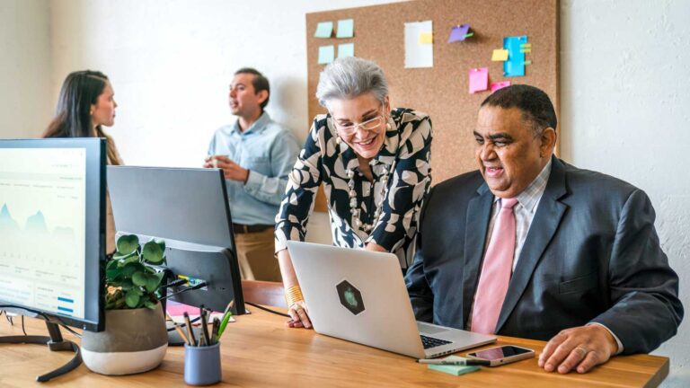 Four colleagues chat at a desk while two examine info on a laptop screen.