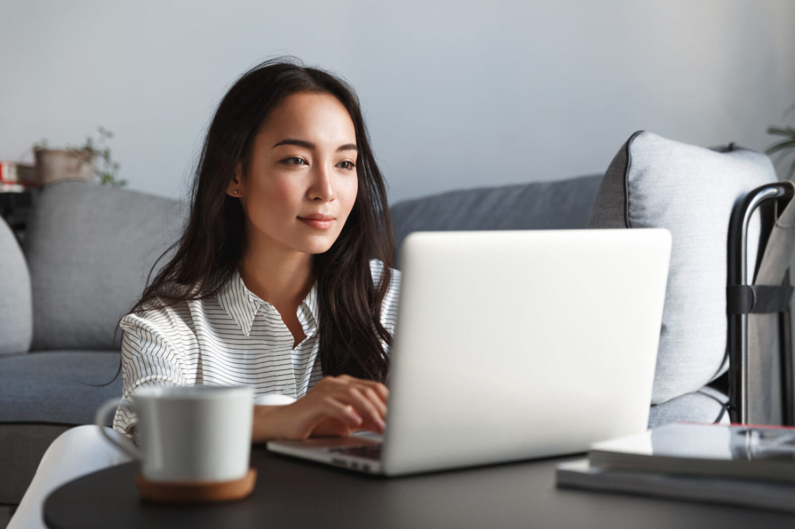 Young ambitious asian girl working remote from home, looking at laptop screen and smiling. Woman checking mail or researching while telecommuting, sitting on floor at her apartment.